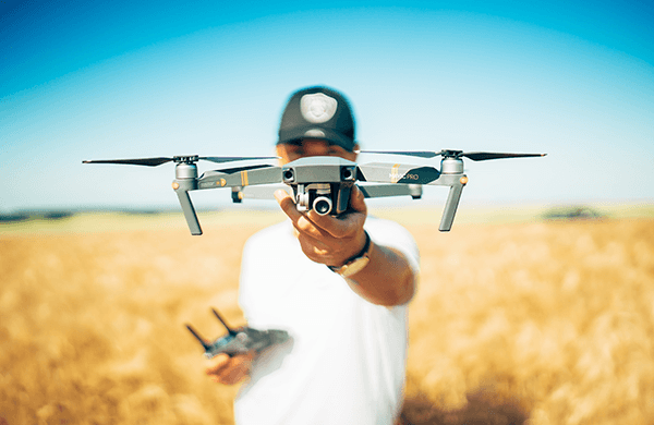 A man in a white shirt and baseball cap holds a drone out in front of his face.