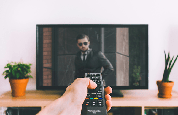 Hand holding remote in front of a television screen displaying man in suit.
