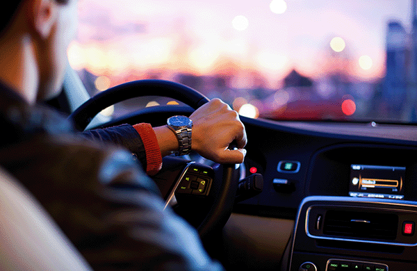 A man with his hand on a steering wheel driving through a city at sunset.