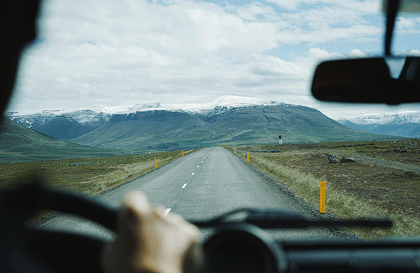 View from behind a person driving down a straight road toward snow capped mountains