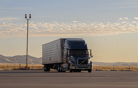 A black semi truck with a white trailer parked in a lot at dusk, mountains in the background.