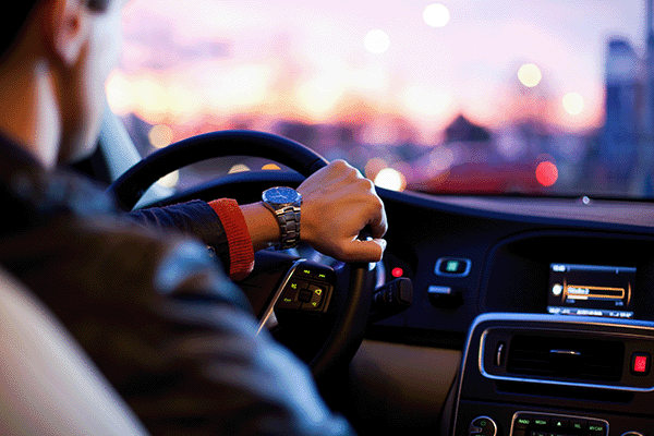 A man with his hand on a steering wheel driving through a city at sunset.